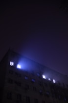 A multi-story building with a dark facade is partially illuminated by lights emanating from several windows. The scene appears to be at night with a mist or fog adding a mysterious glow around the lit windows.