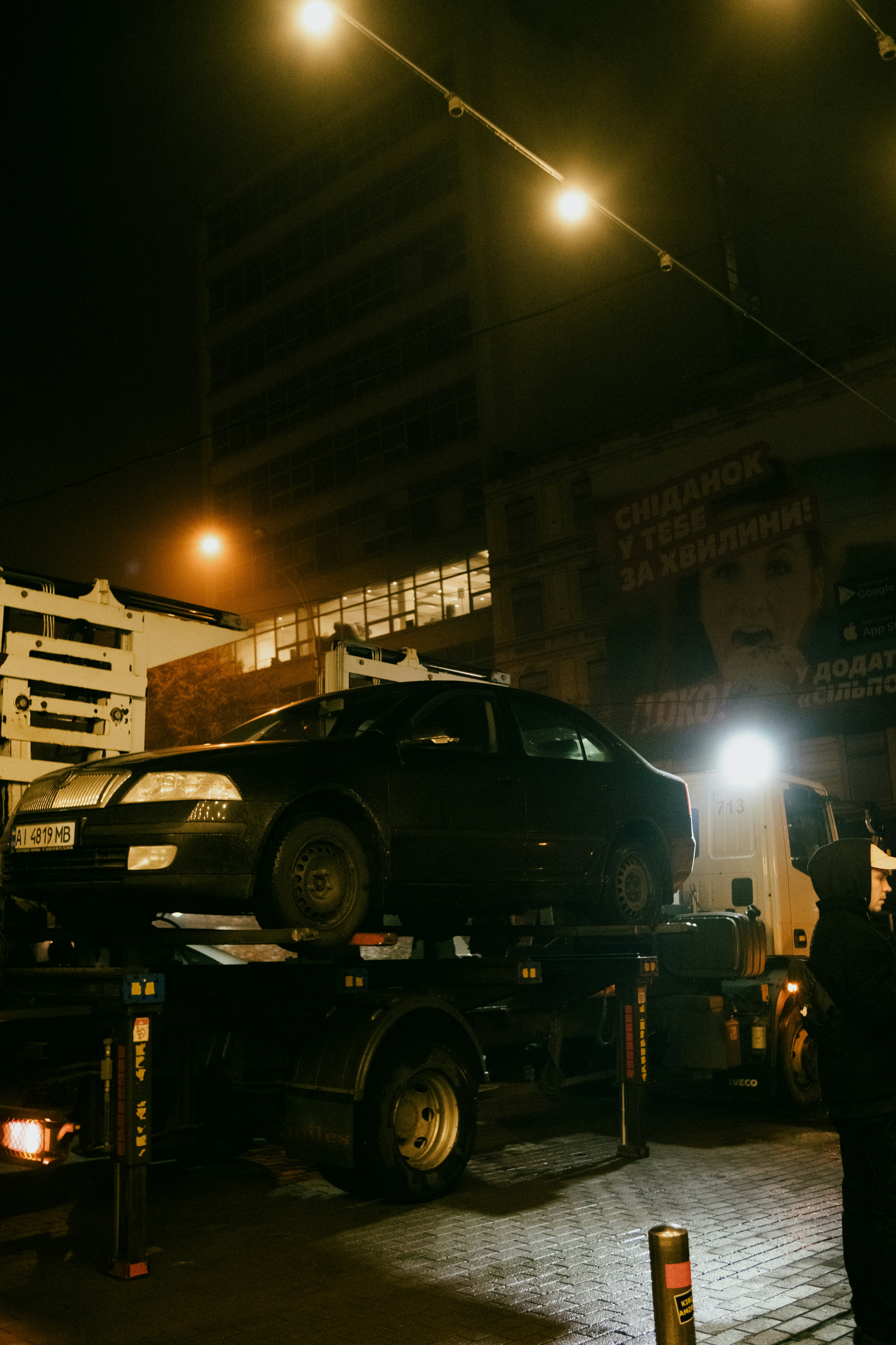Tow truck carrying black sedan at night in urban area with street lights