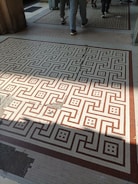 A tiled floor with a geometric pattern of interlocking shapes in brown and beige colors. Several people are walking across the floor, and there is a suitcase on wheels visible. The floor has some areas of wear and damage, with a shadow partially covering it.