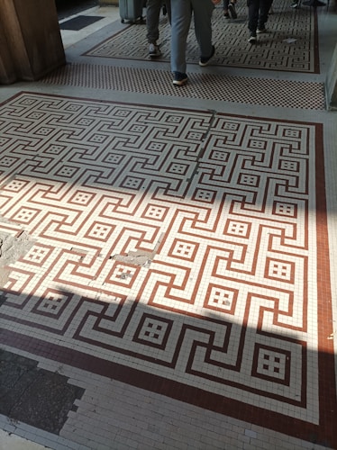 A tiled floor with a geometric pattern of interlocking shapes in brown and beige colors. Several people are walking across the floor, and there is a suitcase on wheels visible. The floor has some areas of wear and damage, with a shadow partially covering it.