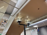 The image captures an indoor setting with a ceiling made of beige-colored panels. Two modern cube-shaped ceiling lights are prominently visible. A net is installed above the ceiling, which may serve as a safety feature. In the background, a snowman decoration with a hat is partially visible, adding a playful element to the environment. Closed metal shutters cover the walls on one side, and a digital sign is attached to the wall.