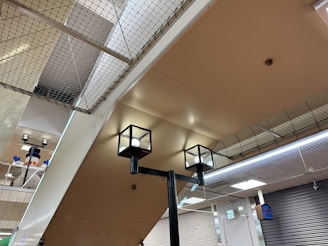 The image captures an indoor setting with a ceiling made of beige-colored panels. Two modern cube-shaped ceiling lights are prominently visible. A net is installed above the ceiling, which may serve as a safety feature. In the background, a snowman decoration with a hat is partially visible, adding a playful element to the environment. Closed metal shutters cover the walls on one side, and a digital sign is attached to the wall.