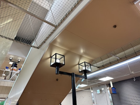 The image captures an indoor setting with a ceiling made of beige-colored panels. Two modern cube-shaped ceiling lights are prominently visible. A net is installed above the ceiling, which may serve as a safety feature. In the background, a snowman decoration with a hat is partially visible, adding a playful element to the environment. Closed metal shutters cover the walls on one side, and a digital sign is attached to the wall.