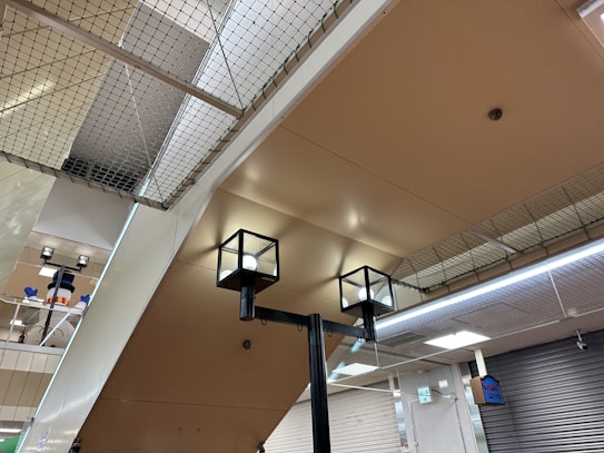 The image captures an indoor setting with a ceiling made of beige-colored panels. Two modern cube-shaped ceiling lights are prominently visible. A net is installed above the ceiling, which may serve as a safety feature. In the background, a snowman decoration with a hat is partially visible, adding a playful element to the environment. Closed metal shutters cover the walls on one side, and a digital sign is attached to the wall.