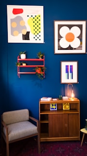 A cozy living space features a deep blue wall adorned with abstract art. The artworks exhibit bold geometric shapes and vibrant colors. A small red shelf holding potted plants is mounted on the wall. Below, a wooden cabinet is topped with books, decorative pieces, and a lamp with an exposed bulb. To the left, a gray upholstered chair provides seating. The overall aesthetic combines modern art with mid-century furniture.