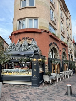 An outdoor café or restaurant setup with a pastry and gelato display case at the corner of a multi-story building. The building features multiple windows with small balconies. The café's decor includes artificial snow-covered trees and greenery, and tables with white chairs are set up for outdoor dining. The building has a rustic aesthetic with stone and warm-colored stucco.