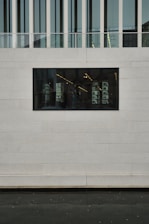 A modern building facade with a rectangular window showing an interior with shelves and lights. The wall is constructed from large concrete or stone slabs, and the upper portion features vertical glass panels and a railing.