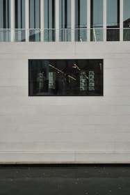 A modern building facade with a rectangular window showing an interior with shelves and lights. The wall is constructed from large concrete or stone slabs, and the upper portion features vertical glass panels and a railing.