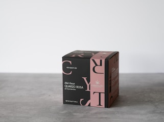 A cube-shaped product box with elegant black and pink design elements, featuring text and logos related to tea. The box is placed on a simple gray surface with a minimalist background.