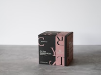 A cube-shaped product box with elegant black and pink design elements, featuring text and logos related to tea. The box is placed on a simple gray surface with a minimalist background.