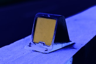 A reflective road marker sits atop a concrete barrier, painted in hues of vivid blue. The marker itself is square-shaped with a yellow reflective surface, contrasting with the deep blue lighting around it.