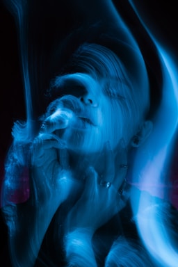A person appears to be immersed in swirling patterns of blue light, with hands near the face. The image has an ethereal and surreal quality created by the long exposure or digital effect.