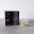 A box labeled 'Crystal Tea Elixir Detox Agata' is prominently displayed, featuring a sleek design with large overlapping text and a small logo of a leaf. Beside the box, there is a transparent pyramid-shaped tea bag containing a variety of loose leaf herbs.