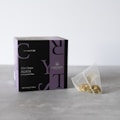 A box labeled 'Crystal Tea Elixir Detox Agata' is prominently displayed, featuring a sleek design with large overlapping text and a small logo of a leaf. Beside the box, there is a transparent pyramid-shaped tea bag containing a variety of loose leaf herbs.
