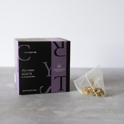 A box labeled 'Crystal Tea Elixir Detox Agata' is prominently displayed, featuring a sleek design with large overlapping text and a small logo of a leaf. Beside the box, there is a transparent pyramid-shaped tea bag containing a variety of loose leaf herbs.