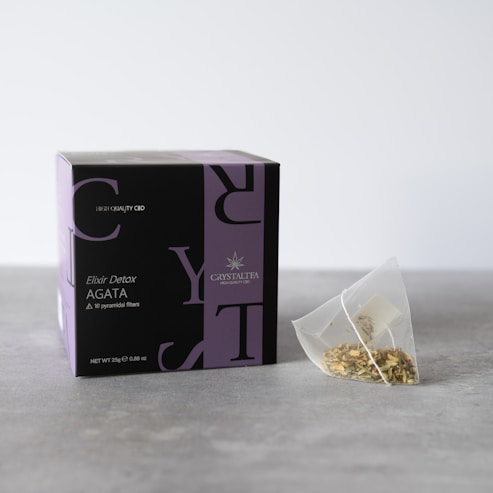 A box labeled 'Crystal Tea Elixir Detox Agata' is prominently displayed, featuring a sleek design with large overlapping text and a small logo of a leaf. Beside the box, there is a transparent pyramid-shaped tea bag containing a variety of loose leaf herbs.