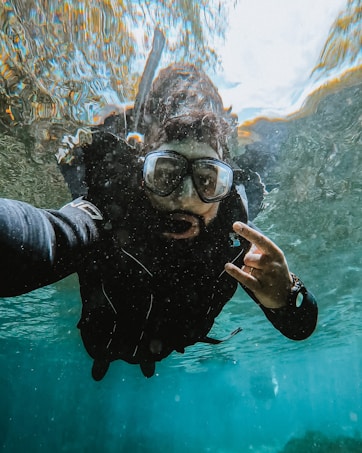 A person is underwater wearing a diving suit and goggles, capturing a selfie. The water is clear, and there is a rippling effect on the surface. The person's hand is making a gesture, possibly a sign or signal.