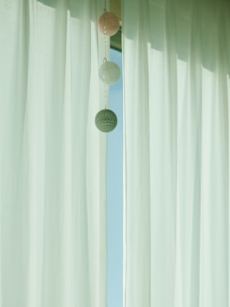 Sheer white curtains are hanging, allowing soft natural light to filter through. Three decorative round balls are strung vertically, adding a playful yet subtle element to the minimalist scene.