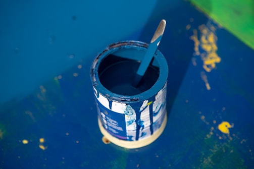 A partially used can of blue paint is open with a paint stick resting inside. The can is on a surface that has splatters of paint and a larger painted area, mostly in shades of blue with hints of yellow and green. The scene suggests a painting or renovation project in progress.