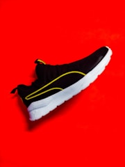 A stylish black sneaker with a white sole is highlighted against a vibrant red background. The shoe features yellow accents along its design and branding on the tongue.