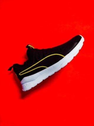 A stylish black sneaker with a white sole is highlighted against a vibrant red background. The shoe features yellow accents along its design and branding on the tongue.