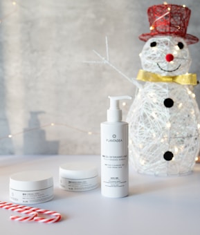 A festive setup with a wire snowman adorned with a red top hat, black buttons, and a golden bow tie on the right side. In the foreground, there are skincare products with white packaging prominently displayed, including a pump bottle and two jars. Two red and white candy canes are placed on a white surface, enhancing the holiday theme. The background is softly illuminated with delicate string lights.