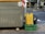 A large gray recycling bin is placed on a paved urban street. Next to it sits a green crate holding a yellow and black insulated delivery bag with a brand logo. The street is lined with trees and bushes, and a road is visible in the background.