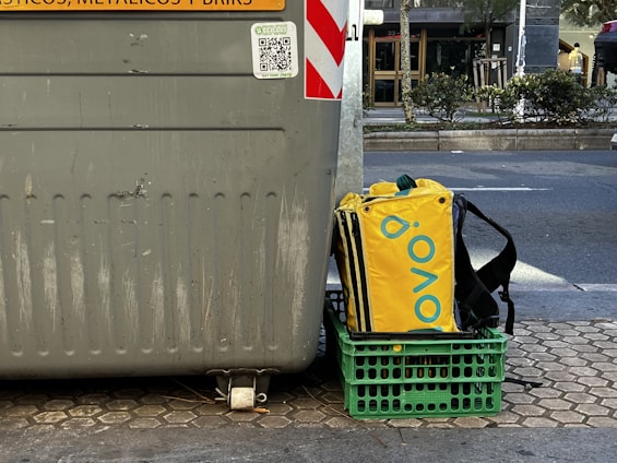A large gray recycling bin is placed on a paved urban street. Next to it sits a green crate holding a yellow and black insulated delivery bag with a brand logo. The street is lined with trees and bushes, and a road is visible in the background.
