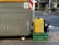 A large gray recycling bin is placed on a paved urban street. Next to it sits a green crate holding a yellow and black insulated delivery bag with a brand logo. The street is lined with trees and bushes, and a road is visible in the background.