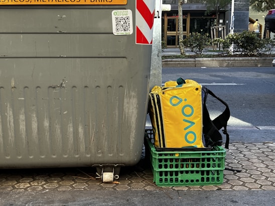 A large gray recycling bin is placed on a paved urban street. Next to it sits a green crate holding a yellow and black insulated delivery bag with a brand logo. The street is lined with trees and bushes, and a road is visible in the background.
