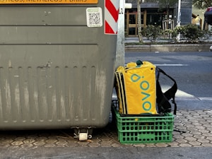 A large gray recycling bin is placed on a paved urban street. Next to it sits a green crate holding a yellow and black insulated delivery bag with a brand logo. The street is lined with trees and bushes, and a road is visible in the background.