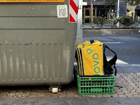 A large gray recycling bin is placed on a paved urban street. Next to it sits a green crate holding a yellow and black insulated delivery bag with a brand logo. The street is lined with trees and bushes, and a road is visible in the background.