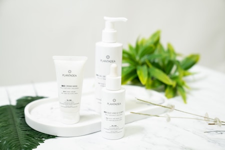 Three skincare product bottles are displayed on a white circular tray. The background includes fresh green leaves, emphasizing a natural theme. The surface is a marble-like white, adding elegance.