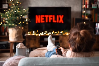 A person is relaxing on a sofa, facing a TV screen displaying the Netflix logo. In the foreground, a hand holds a remote control. A mug with a hot drink topped with whipped cream is placed nearby on a table. A decorated Christmas tree with lights adds a festive touch to the background.