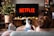 A person is relaxing on a sofa, facing a TV screen displaying the Netflix logo. In the foreground, a hand holds a remote control. A mug with a hot drink topped with whipped cream is placed nearby on a table. A decorated Christmas tree with lights adds a festive touch to the background.