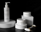 A sleek arrangement of skincare products including a pump bottle and two jars, all labeled with the brand 'PlantaDea'. These products are placed on geometric white platforms against a dark background, accompanied by a marble-textured facial roller with gold accents.