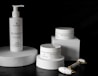 A sleek arrangement of skincare products including a pump bottle and two jars, all labeled with the brand 'PlantaDea'. These products are placed on geometric white platforms against a dark background, accompanied by a marble-textured facial roller with gold accents.