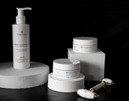 A sleek arrangement of skincare products including a pump bottle and two jars, all labeled with the brand 'PlantaDea'. These products are placed on geometric white platforms against a dark background, accompanied by a marble-textured facial roller with gold accents.