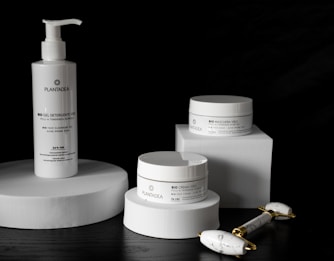 A sleek arrangement of skincare products including a pump bottle and two jars, all labeled with the brand 'PlantaDea'. These products are placed on geometric white platforms against a dark background, accompanied by a marble-textured facial roller with gold accents.