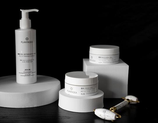 A sleek arrangement of skincare products including a pump bottle and two jars, all labeled with the brand 'PlantaDea'. These products are placed on geometric white platforms against a dark background, accompanied by a marble-textured facial roller with gold accents.