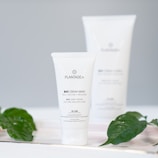 A tube of Plantadea bio hand cream is positioned in the foreground, with clear labeling in both Italian and English. The product is marketed as suitable for dry and delicate skin and contains 1% CBD. The background is slightly blurred, featuring a larger version of the product. Green leaves are artfully placed around the product, adding an organic touch.