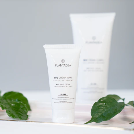 A tube of Plantadea bio hand cream is positioned in the foreground, with clear labeling in both Italian and English. The product is marketed as suitable for dry and delicate skin and contains 1% CBD. The background is slightly blurred, featuring a larger version of the product. Green leaves are artfully placed around the product, adding an organic touch.