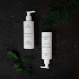 Two white bottles with pump dispensers are placed on a dark textured surface. The bottles are labeled Plantadea and are likely skincare products. Green leaves with water droplets are positioned around the bottles, adding a natural element to the composition.