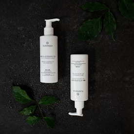 Two white bottles with pump dispensers are placed on a dark textured surface. The bottles are labeled Plantadea and are likely skincare products. Green leaves with water droplets are positioned around the bottles, adding a natural element to the composition.