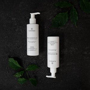 Two white bottles with pump dispensers are placed on a dark textured surface. The bottles are labeled Plantadea and are likely skincare products. Green leaves with water droplets are positioned around the bottles, adding a natural element to the composition.