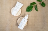 Two skincare products, a tube and a dropper bottle, are placed on wooden slabs. The background is made of burlap fabric, and a few green leaves are positioned to the upper right.