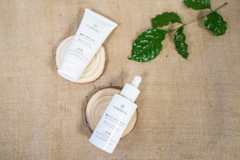 Two skincare products, a tube and a dropper bottle, are placed on wooden slabs. The background is made of burlap fabric, and a few green leaves are positioned to the upper right.