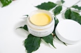 A jar of face and body ointment with a creamy yellow substance inside is placed on a white surface. The jar is open, and the lid is lying next to it. Green leaves form a natural backdrop, adding an organic touch.