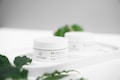A white jar of bio face cream with the label 'PLANTADEA' is placed on a tray. The scene is minimalist and bright, with a soft focus background. Green leaves are visible, adding a natural element to the image.