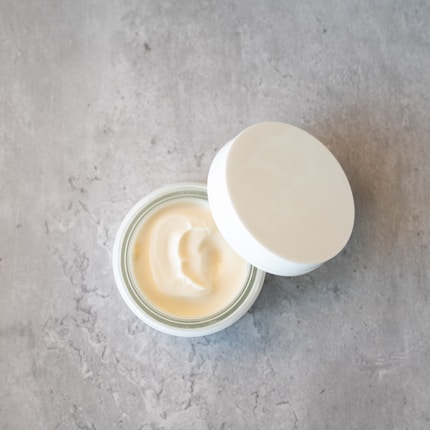 A container with an open lid, showing a creamy, off-white substance inside. The container is placed on a flat, gray stone or concrete surface, providing a minimal and clean aesthetic.